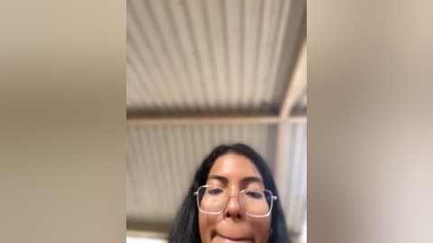 Media: A video of a smiling, light-skinned woman with long black hair, wearing glasses, standing indoors with a corrugated metal ceiling. The background is blurred, focusing on her face.