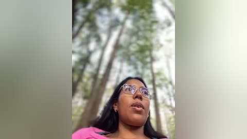 Media: A video of a young woman with medium brown skin, long black hair, and round glasses, standing in a lush, sunlit forest with tall trees and green foliage in the background.