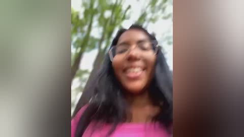 Media: A blurry video of a smiling Black woman with long black hair and wearing a pink top, standing outdoors with green foliage in the background.