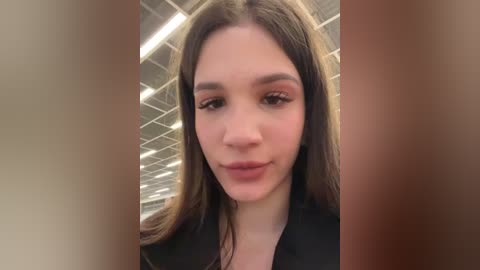 Media: Video of a young woman with light skin, long brown hair, wearing a black top, indoors with fluorescent lights and white ceiling tiles.