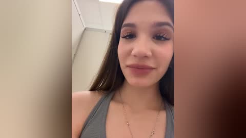 Media: A close-up video of a young woman with long dark hair, wearing a gray tank top, standing in a room with beige walls and a white ceiling.