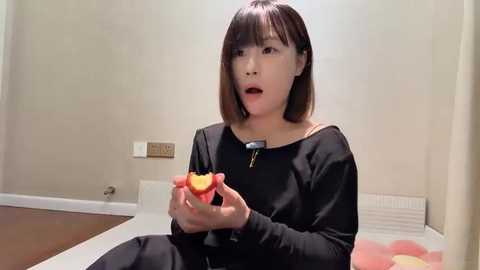 Media: Video of a young Asian woman with straight brown hair, wearing a black sweater, holding a peeled orange in a beige room with a wooden floor.