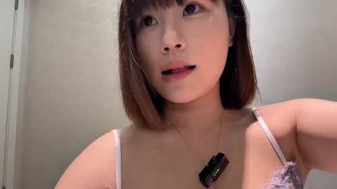 Media: Video of an East Asian woman with straight, shoulder-length brown hair, wearing a white lace bra, and a necklace with a black box pendant. She gazes upwards, lips slightly parted. Background is a plain, light-colored wall.