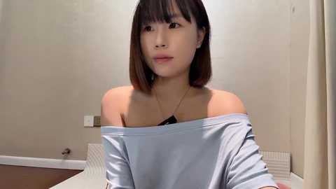 Media: Video of an East Asian woman with straight, shoulder-length brown hair, wearing a loose, off-shoulder light blue shirt, in a minimalist room with beige walls and a radiator.