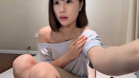 Media: Video of an Asian woman with straight brown hair, wearing a light grey off-shoulder top, sitting indoors on a white surface, looking directly at the camera.