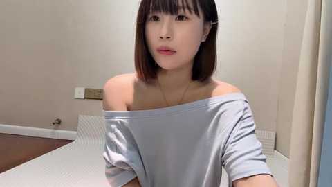 Media: Video of a young Asian woman with shoulder-length black hair and fair skin, wearing a light blue off-shoulder top, seated in a minimalist room with beige walls and white furniture.