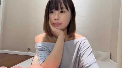 Media: Video of a young East Asian woman with straight, shoulder-length black hair, wearing a light blue off-shoulder top, sitting indoors with a contemplative expression, hand on chin, against a plain beige wall and wooden floor.