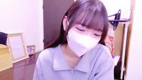 Media: Video of a young Asian woman with long dark hair, wearing a white surgical mask and light blue shirt, standing indoors, with a wooden chair and framed artwork in the background.