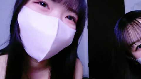 Media: Video of an East Asian woman with long black hair, wearing a white medical mask and a black top, looking directly at the camera.