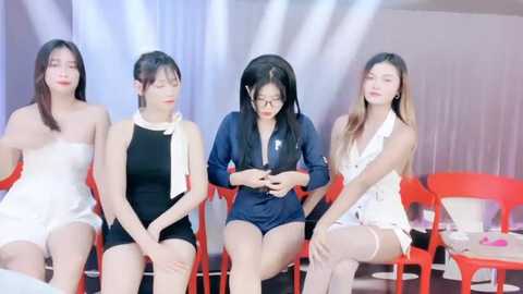 Media: Video of five Asian women in various outfits, including a white strapless dress, a black one-piece swimsuit, a navy blue romper, a white lace romper, and a black dress, sitting on red chairs against a white backdrop.