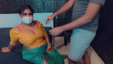 Media: Video of a woman in a yellow top, green sari, and face mask, being pushed by a man in blue shorts in a dimly lit room.