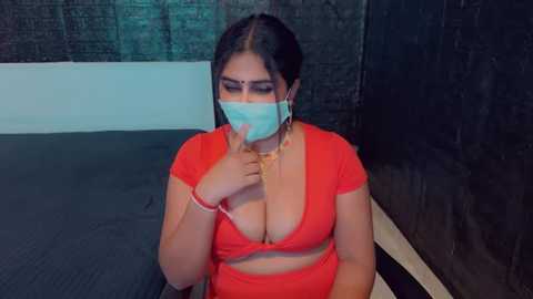 Media: Video of a South Asian woman with dark skin, wearing a blue surgical mask, red crop top revealing cleavage, and red pants, seated on a bed with a dark curtain backdrop.