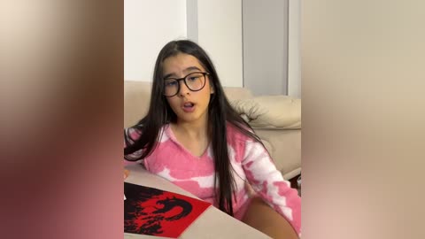 Media: Video of a young woman with long dark hair, wearing black-framed glasses and a pink tie-dye shirt, seated on a beige couch, holding a book with red and black cover, indoors.