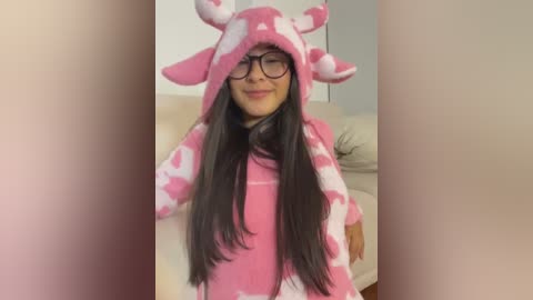 Media: Video of a smiling woman with long black hair, wearing a pink cow onesie with white spots, glasses, and black boots, standing indoors with a beige couch in the background.