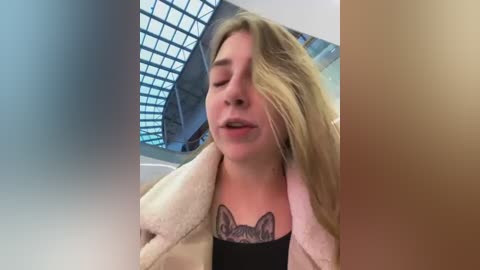 Media: Video of a blonde woman with closed eyes, wearing a beige coat over a black top, showcasing a tattoo of a fox head on her chest. The background features a modern, glass-roofed atrium.