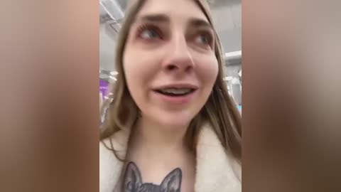 Media: Video of a smiling young woman with light skin, light brown hair, and a gray cat tattoo on her chest, in a modern retail store with shelves and fluorescent lights in the background.