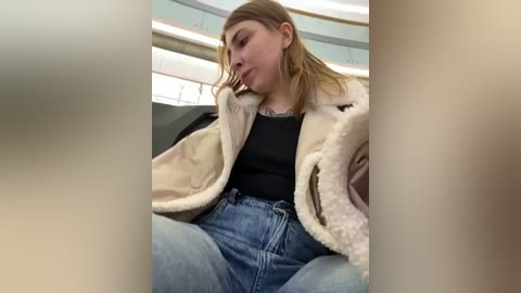 Media: Video of a young, fair-skinned woman with light brown hair, wearing a beige fur-trimmed coat over a black top and blue jeans, sitting in a modern, circular seating area.