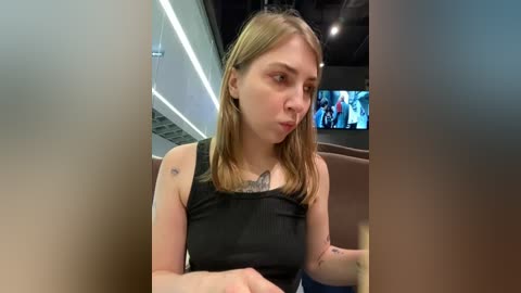 Media: Video of a young Caucasian woman with straight, shoulder-length blonde hair, wearing a black tank top, sitting indoors with a surprised expression. Background includes a TV screen and modern decor.