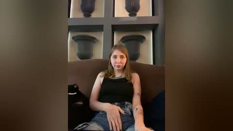 Media: Video of a young, fair-skinned, slim woman with shoulder-length brown hair, wearing a black tank top and high-waisted jeans, sitting on a brown couch, surrounded by black decorative vases on shelves in a dimly lit room.