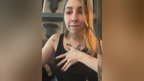 Media: Video of a young woman with light skin, long blonde hair, wearing a black tank top, sitting indoors with a cat. She has tattoos on her shoulders and fingers.