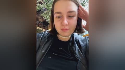 Media: Video of a young woman with shoulder-length brown hair, wearing a black leather jacket and choker, looking down, with blurred greenery and building in the background.