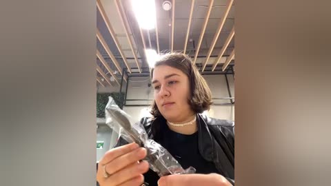 Media: Video of a young woman with shoulder-length dark hair, wearing a black leather jacket and a choker, examining a plastic-wrapped object. She is indoors with a ceiling of exposed wooden beams and fluorescent lights.