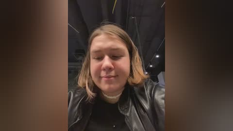 Media: A video of a young Caucasian woman with light skin, shoulder-length blonde hair, wearing a black leather jacket and a white turtleneck, smiling softly in a dimly lit indoor setting.