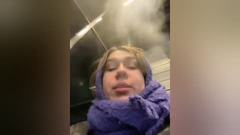 Media: Video of a young woman with light skin and brown hair, wrapped in a purple towel, in a steamy bathroom with white tiles and a fogged mirror.