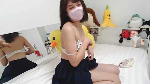 Media: A video of an East Asian woman with straight black hair, wearing a white face mask, navy blue skirt, and white bra, adjusting her bra in a room filled with plush toys and a bed.