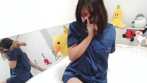 Media: A video shows a woman with shoulder-length brown hair, wearing a blue dress, sitting on a bed, using a mirror to apply makeup. Background features a white wall with yellow duck and cat stickers.