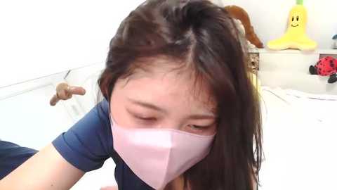 Media: Video of an Asian woman with long brown hair, wearing a pink face mask, leaning over a table. Background includes a white wall, a stuffed yellow bear, and a red toy.