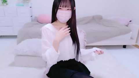 Media: Video of an Asian woman with long black hair, pale skin, wearing a white long-sleeve top, black skirt, and pink face mask, seated on a white rug in a minimalist bedroom with a white dresser and bed.