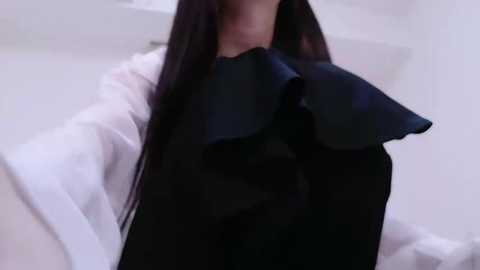 Media: Video of a person with long, straight black hair, wearing a white shirt and a large, black ruffled bow tie. The background is a blurred, light-colored room with minimalistic decor.