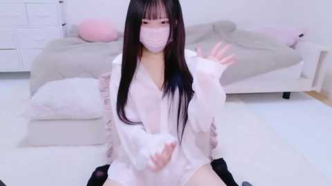 Media: A video of an Asian woman with long black hair, wearing a white surgical mask and an oversized white shirt, sitting on the floor in a modern bedroom with white furniture.