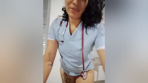 Media: Video of a curvaceous, light-skinned woman with curly black hair wearing a light blue nurse uniform and red stethoscope, standing in a clinical room with blinds.