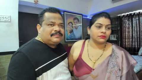 Media: Video of an elderly South Asian couple in a living room, wearing traditional attire; man in black and white shirt, woman in maroon sari with gold necklace.