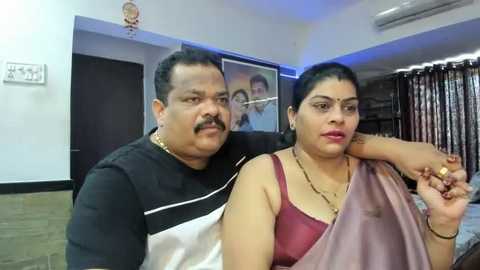 Media: Video of an Indian couple in a living room. The man, with a mustache, wears a black shirt, while the woman, with short black hair, sports a maroon saree. Background shows a framed photo, curtains, and a clock.