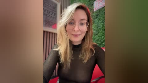 Media: A video of a fair-skinned, blonde woman with glasses, wearing a sheer black top, sitting in a red chair against a green wall.