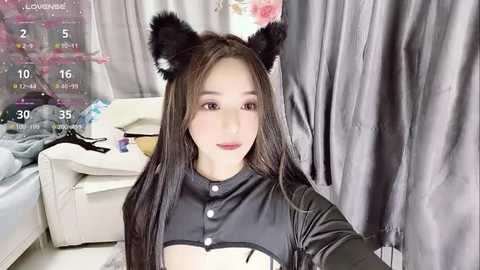 Media: A young East Asian woman with long brown hair and cat ear headband poses in a modern bedroom, wearing a black leather-like top, against gray curtains.