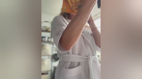 Media: A video of a woman in a light grey, textured robe, holding an orange book, taken in a bright, modern kitchen with white cabinets and shelves.