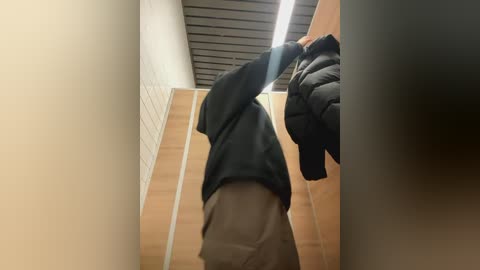 Media: Video of a person wearing a black jacket and light brown pants, standing in a narrow, fluorescent-lit hallway with white tiled walls and a dark wooden ceiling.