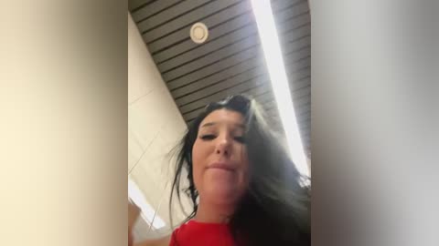 Media: Video of a woman with long black hair, wearing a red top, standing in a modern room with a slanted ceiling and fluorescent lighting.