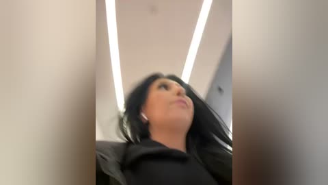 Media: A low-angle video of a woman with long black hair, wearing a black jacket, looking upward, standing in a modern indoor setting with bright overhead lights.