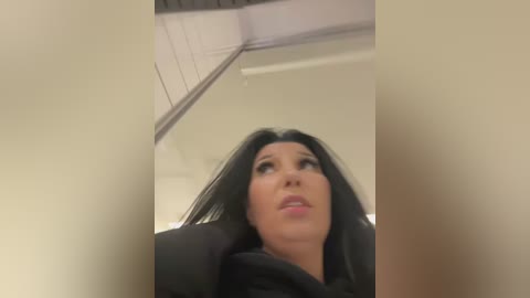 Media: Video of a woman with long, black hair and fair skin, wearing a black top, looking up with an expression of surprise or shock. The background features a white ceiling and part of a wall.