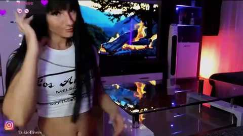 Media: Video of a slender, light-skinned woman with long black hair, wearing a white crop top with \"Los Angeles\" text, standing in a dimly lit living room with a TV showing a mountain fire scene, a glass table, and a black chair.