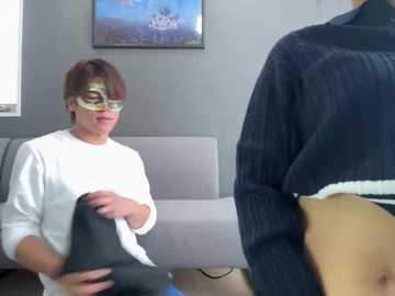 Media: Video of a person in a white shirt and a gold mask standing next to a person in a black sweater, in a modern living room with a gray couch and a framed painting on the wall.