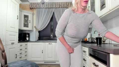 Media: Video of a blonde woman in a sheer, light-gray top, revealing her large breasts and nipples, standing in a modern kitchen with white cabinets and a dark countertop.