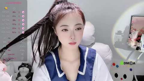 Media: A video of an Asian woman with fair skin and long, straight black hair, wearing a blue sailor collar school uniform, sitting in a bright, modern room with a calendar and a computer screen in the background.