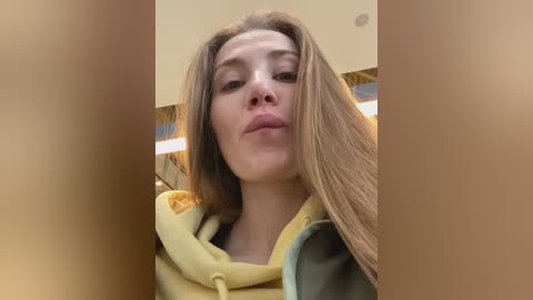 Media: Video of a young Caucasian woman with long, straight brown hair, wearing a yellow hoodie, making a pouty face. Background includes beige walls and a ceiling light.