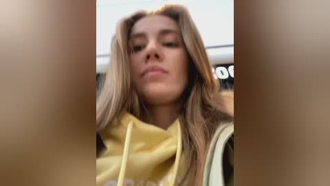 Media: A video of a young woman with long, straight blonde hair, wearing a yellow hoodie, taken from a low angle, with a blurred background.